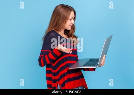 Puzzled woman wearing striped casual style sweater, standing with confused facial expression, holding laptop and looking at display. Indoor studio shot isolated on blue background. Stock Photo