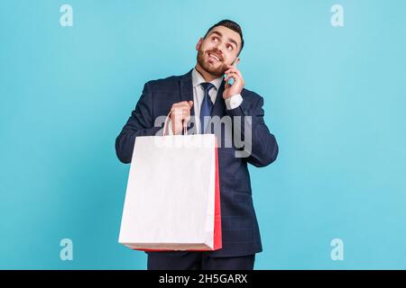 Young adult happy man wearing dark office suit holding shopping bags in hands and talking phone, boasting his purchases with huge discounts. Indoor studio shot isolated on blue background. Stock Photo
