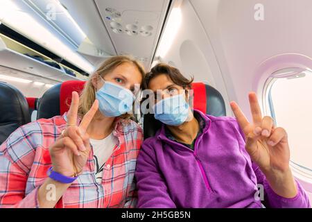 young couple wearing a face mask for COVID-19 protection sitting in an airplane window seat showing two finger V-shaped sign on left hand to the camer Stock Photo