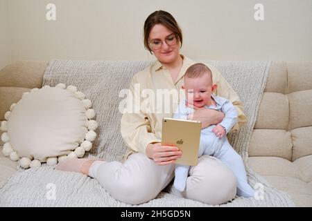 Happy woman mother with infant baby watching in a digital tablet iPad Apple while sitting on the home sofa in the living room - Moscow, Russia, Novemb Stock Photo