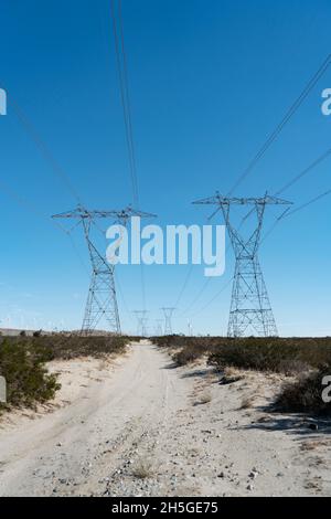 A dirt road following between the path to two high tension power towers and lines stretching across desert. Stock Photo