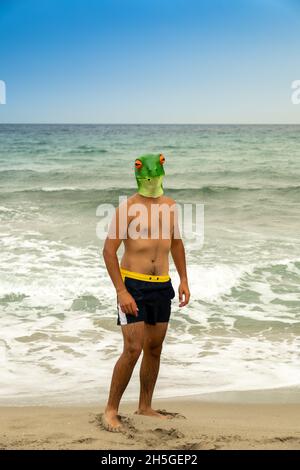 Vertical shot of a Caucasian male with a frog head on the beach Stock Photo