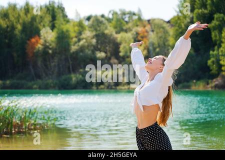 A young woman, against the background of a beautiful forest lake, raised her hands up, depicting happiness. Stock Photo