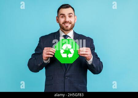 Positive bearded man wearing official style suit holding green recycling sign in hand and looking smiling at camera, thinking green. Indoor studio shot isolated on blue background. Stock Photo