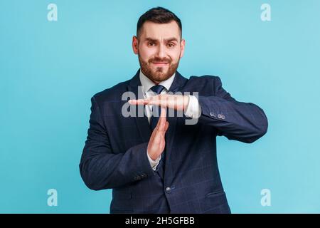 I need more time. Portrait of frustrated brunette man with beard in dark suit showing time out gesture, looking at camera, hurry to meet deadline. Indoor studio shot isolated on blue background. Stock Photo