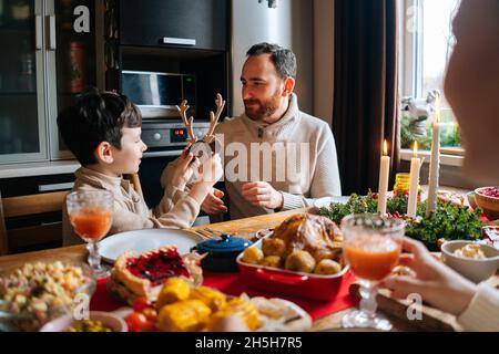 Medium shot of happy father putting toy reindeer antlers on head of little child boy sitting at festive Christmas dinner table. Stock Photo