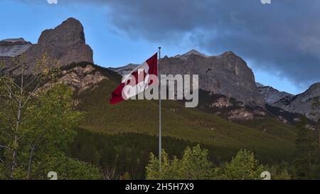 Huge red and white colored Canadian flag flying in the wind between trees in front of majestic mountains near Canmore, Kananaskis Country, Canada. Stock Photo