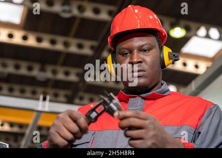 Portrait Of Black Industrial Worker In Red Helmet, Hearing Protectors And Work Uniform. Stock Photo