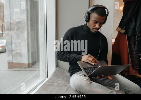 Young black man wearing headphones sitting on a windowsill, attentively looking at laptop screen. On the first floor next to a street. Stock Photo