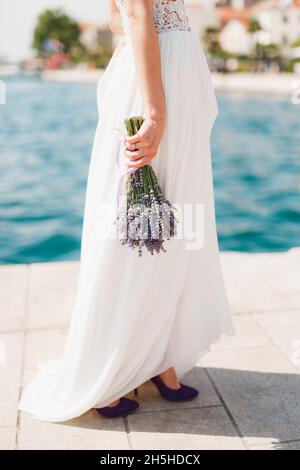 Bride in a white lace dress with a bouquet of lavender in her hands stands on the pier by the sea against the background of the ancient houses of the Stock Photo