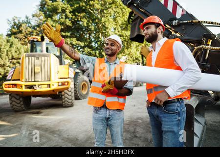 Two men engineers discussing their work standing against construction machines Stock Photo