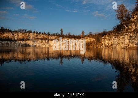 Rock cliffs lit by orange sunset sun light with autumn trees atop reflecting in water, Artificial water reservoir, former limestone quarry in Krakow Stock Photo