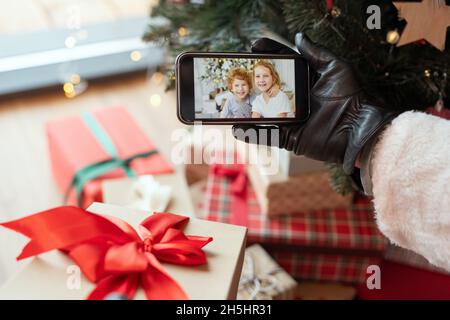 Hand in black leather glove holding smartphone with photo of kids on screen against Christmas presents and tree Stock Photo