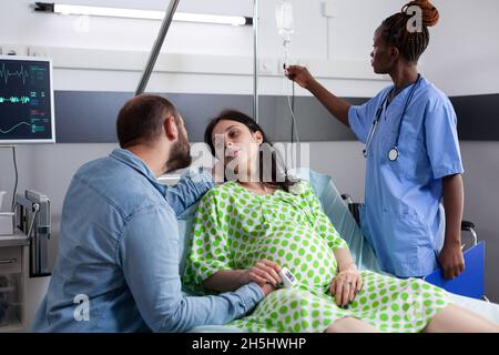 Couple with pregnancy waiting on child delivery in hospital ward at clinic. African american nurse checking IV drip bag while pregnant woman laying in bed and man comforting about childbirth Stock Photo