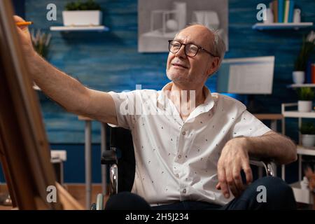 Portrait of disabled artist using pencil on canvas for professional drawing in workshop room. Caucasian old man with handicap working on modern creation while sitting in wheelchair Stock Photo