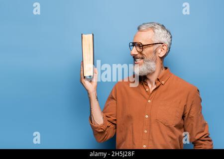 tattooed middle aged teacher in glasses smiling and holding book on blue Stock Photo