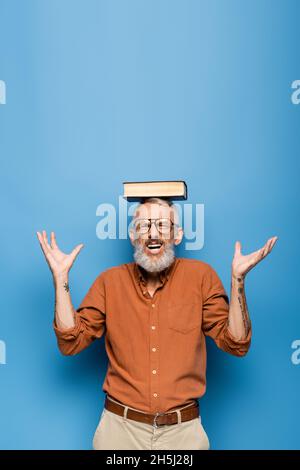 tattooed middle aged teacher in glasses and book on head gesturing on blue Stock Photo