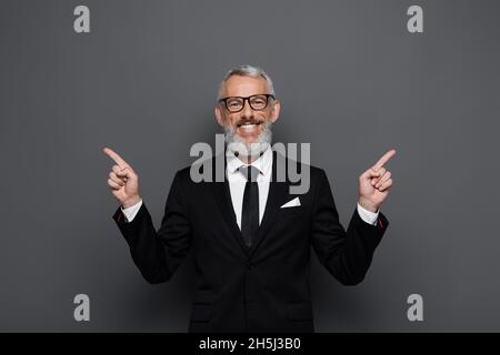 positive middle aged businessman in glasses pointing with fingers aside on grey Stock Photo