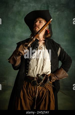 Cropped portrait of brutal man, medeival pirate in vintage costume with pistol, gun isolated over dark background. Stock Photo