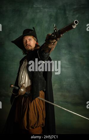 Cropped portrait of brutal frightening man, medeival pirate in vintage costume with sword and gun isolated over dark background. Stock Photo
