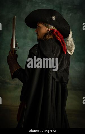 Cropped portrait of brutal frightening man, medeival pirate in vintage costume with sword and gun isolated over dark background. Side view Stock Photo