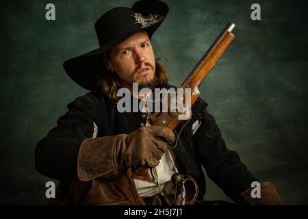 Cropped portrait of brutal frightening man, medeival pirate in vintage costume raising gun sitting isolated over dark background. Stock Photo