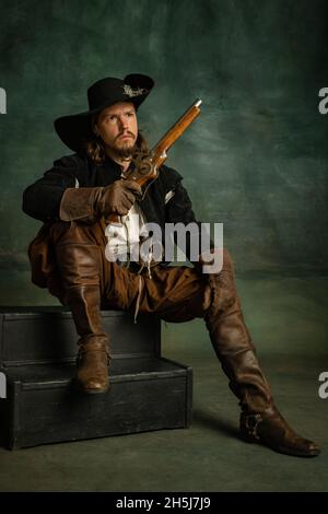 Cropped portrait of brutal frightening man, medeival pirate in vintage costume with sword and gun sitting isolated over dark background. Stock Photo