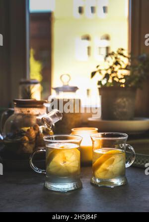 Two glass tea cups with fresh lemon tea on kitchen table with tea pot, herbs and kitchen utensils at window background. Healthy homemade tea with vita Stock Photo