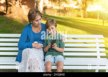 Grandson teaches grandmother how to use cell phone while sitting on park bench. Concept of old people and new technologies Stock Photo