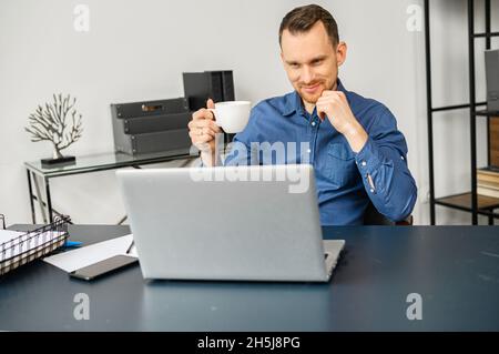 Male freelancer drinking coffee, using a laptop for work in a office. A millennial guy works on outsours, receiving emails, conducts correspondence with customer Stock Photo