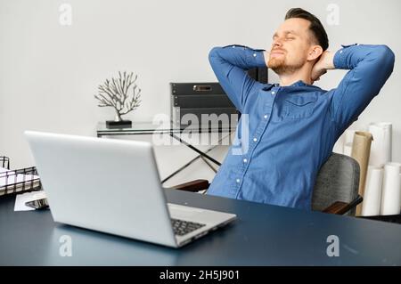 Peaceful millennial guy sitting at the desk in the modern home office, feeling excited for completing the project, placed hands behind head and stretching, enjoying remote freelance job Stock Photo