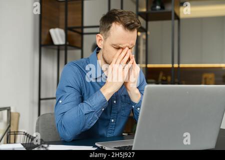 Fatigued guy using laptop for working, a man in smart casual wear is rubs the bridge of the nose, feels tired and burnout, sitting at the desk in modern office with eyes closed Stock Photo