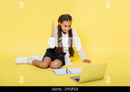 Smiling schoolkid raising hand during video call on laptop near notebook on yellow background Stock Photo