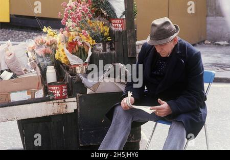 London 1982. A street view of a flower salesman. The tulips and flowers are standing in old gallon beer cans Watneys party seven. Credit Roland Palm. Stock Photo