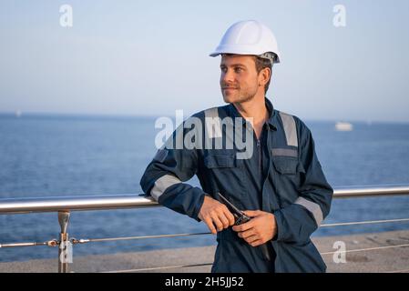 Confident smiling young bearded male worker in protective clothes and hardhat with walkie talkie in hands leaning against metal railing and looking away while having break during work in sea port Stock Photo