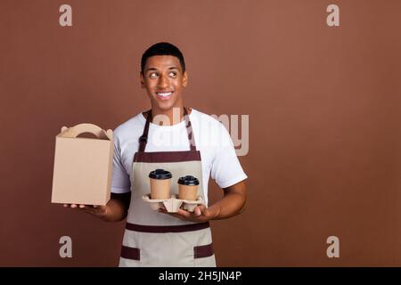 Photo of young afro man worker in coffee bar shop hold paper cup with hot latte beverage isolated over brown color background Stock Photo