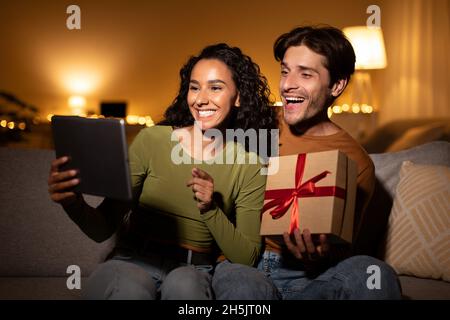 Happy Couple Making Video Call Holding Wrapped Gift Box Congratulating Somebody Celebrating Christmas Online, Using Digital Tablet At Home. Modern Hol Stock Photo