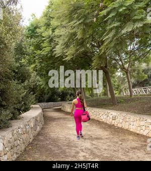Smiling dark haired woman dressed sportswear walking with sports bag in a park, back view. Young caucasian women in pink clothing outdoor. Sport and h Stock Photo