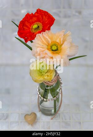 View form above of colorful poppies and ranunculus in a glass vase wrapped with leather cord and a string of rhinestones, next to a heart-shaped stone Stock Photo