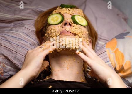 A woman makes a cosmetic mask at home from oatmeal on her face and cucumber. Anti-aging treatments, skin care concept. Stock Photo