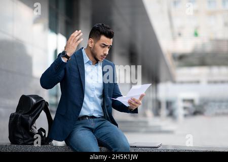 Emotional arabic guy young entrepreneur with backpack sitting on the street next to office building, reading mail and gesturing, businessman got offic Stock Photo