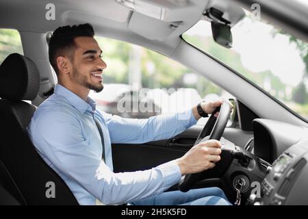 Joyful bearded arabic guy in formal outfit looking at road and smiling, driving nice automobile, young middle eastern businessman going to office, sid Stock Photo