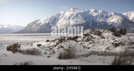 People walking along the rocky outpost of Beluga Point at Turnagain Arm of Cook Inlet in early spring with snow covered mountain landscape and icy ... Stock Photo