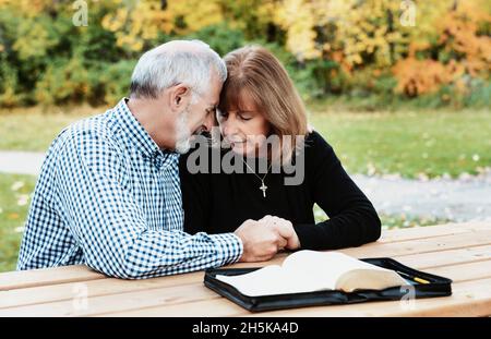 A mature couple sharing devotional time together by studying the Bible and praying together at a picnic table on a warm fall day in a city park Stock Photo