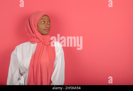 Beautiful young dark-skinned young woman of Arab appearance, looks sideways cheerfully, wears white shirt and pink veil, has healthy skin, in focus is Stock Photo