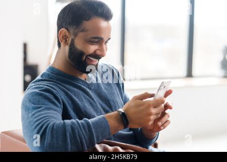 Cheerful hilarious Indian middle-aged man using smartphone sitting on the couch at home, happy eastern guy watching comedy show and laughing, dark-haired hispanic male enjoying online chatting Stock Photo