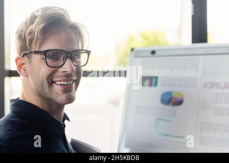 Smiling programmer in eyeglasses looking at camera near blurred flip chart Stock Photo