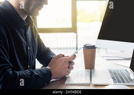 Cropped view of programmer using smartphone near computers and coffee to go in office Stock Photo