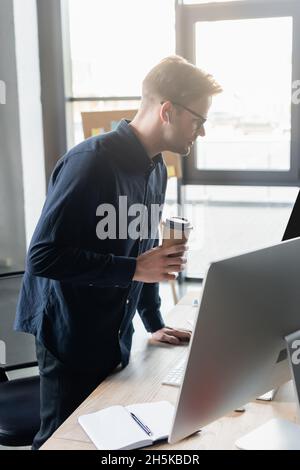 Side view of programmer in earphone holding paper cup near computers and notebook in office Stock Photo