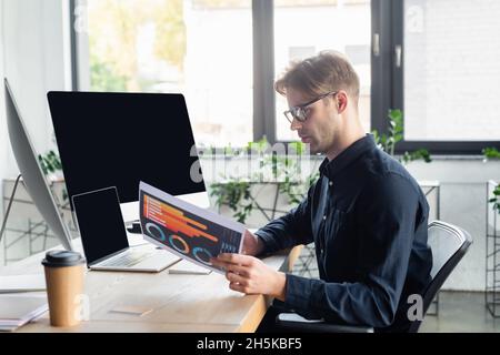 Side view of programmer working with paper near computers and coffee in office Stock Photo